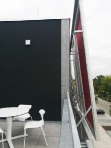 Exterior terrace featuring a white table and chairs next to a black stucco wall, looking towards the steel framework and red backing of the building's facade shading system.