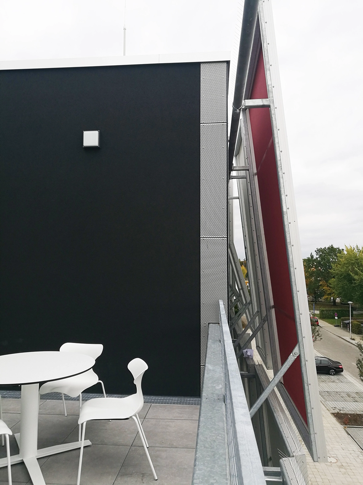 Exterior terrace featuring a white table and chairs next to a black stucco wall, looking towards the steel framework and red backing of the building's facade shading system.