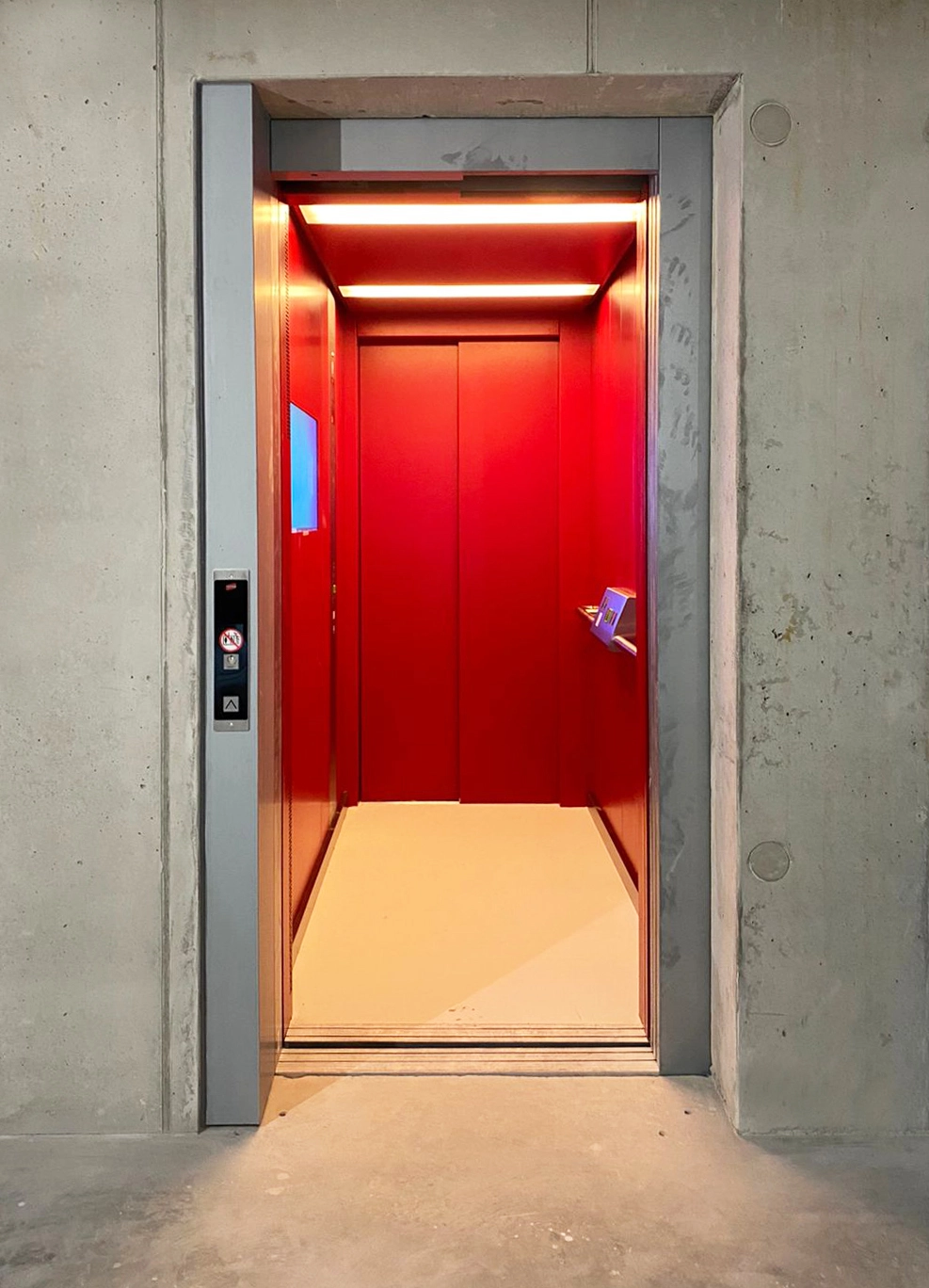 Frontal view of an open elevator cabin with a striking crimson red interior, set within a raw, unfinished concrete structural wall.