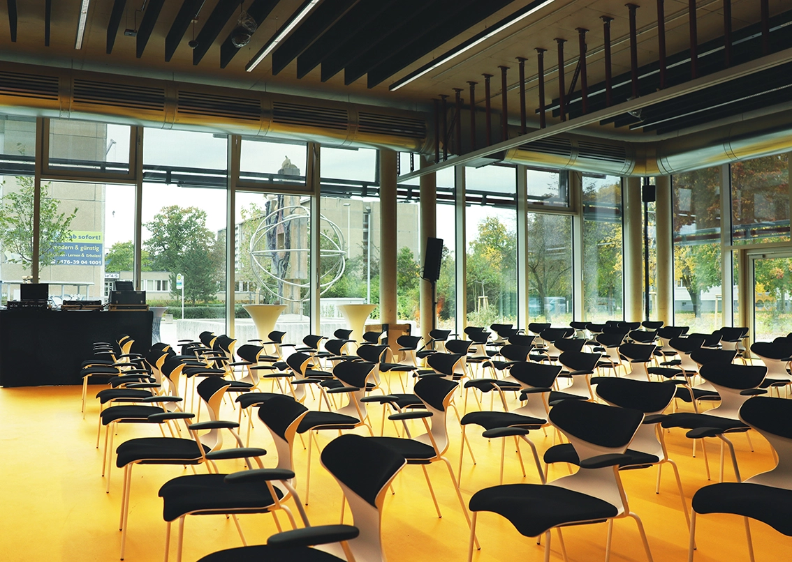 Interior of a modern presentation hall featuring striking yellow flooring, rows of black designer chairs, floor-to-ceiling windows, and exposed industrial ceiling infrastructure.