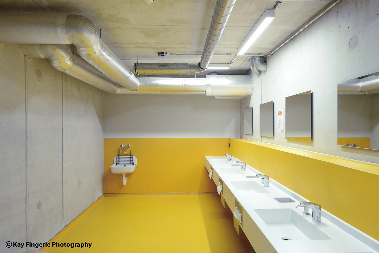 Minimalist public restroom featuring a vibrant yellow floor and matching wall panel, raw concrete surfaces, a row of white sinks, and exposed metallic ceiling pipes.