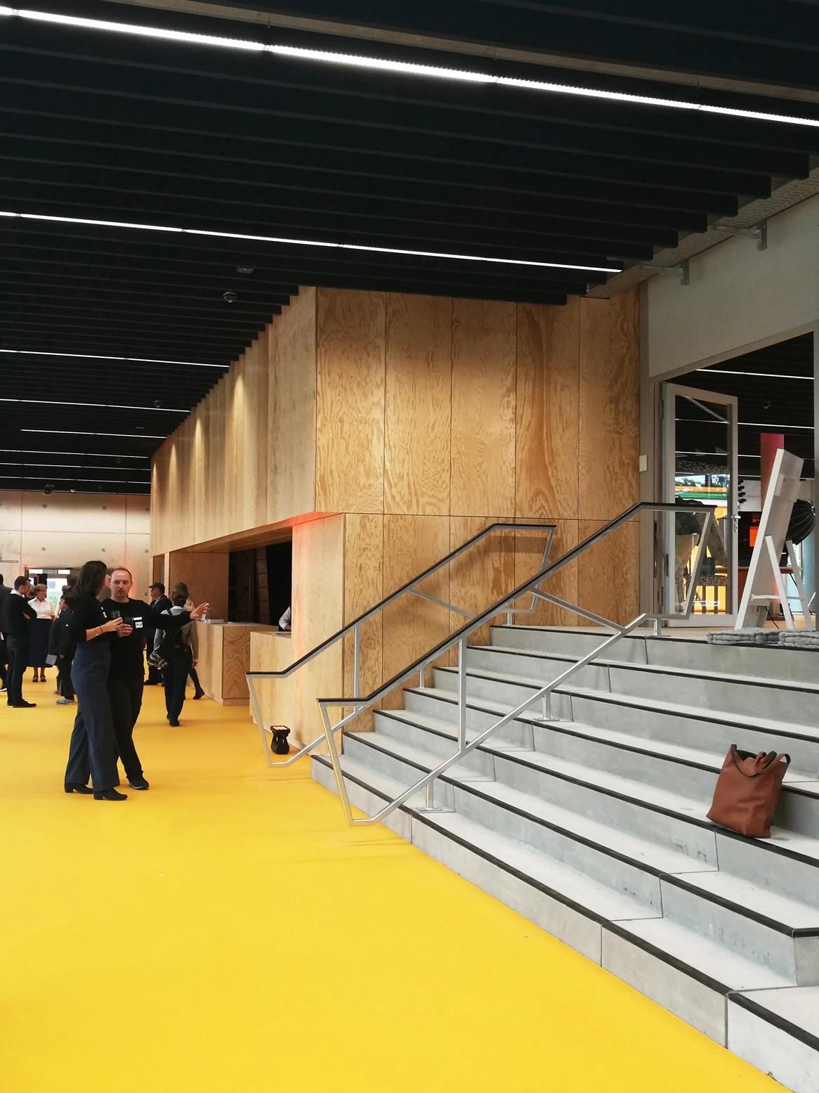 Interior view of the open event space showing wide concrete steps used as informal seating, a matching plywood service hub, and a bright yellow floor.