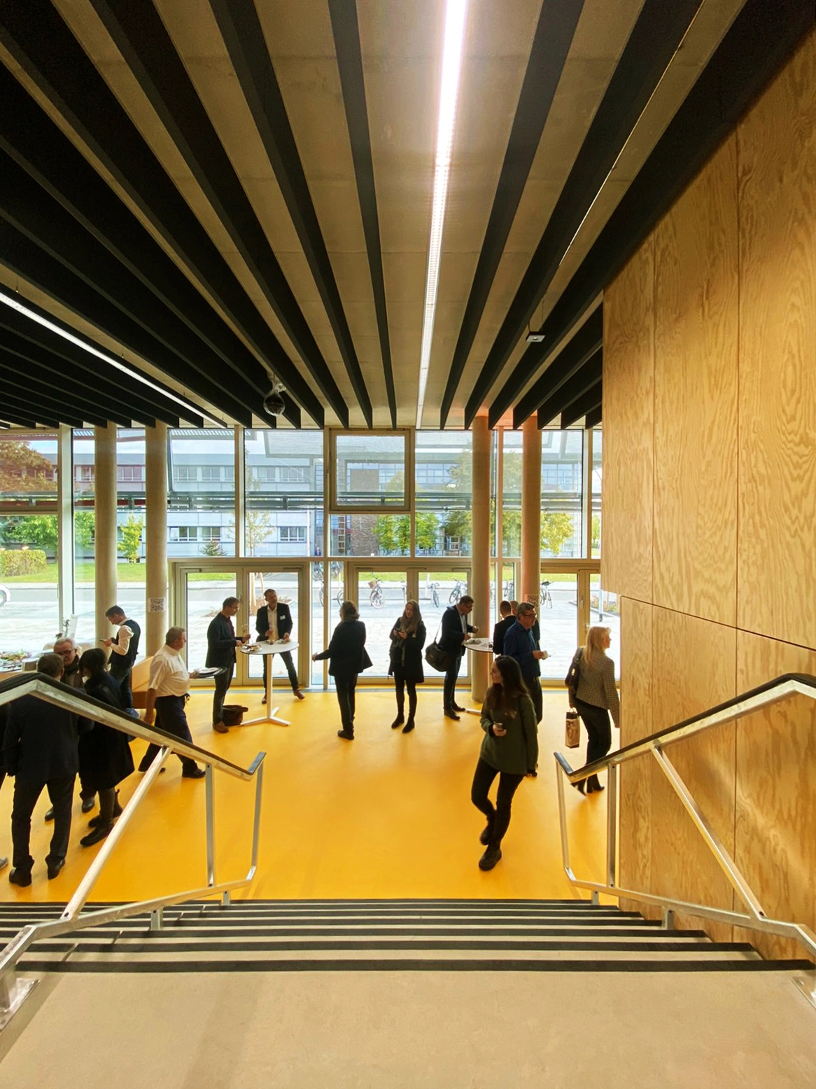 Looking down a concrete staircase with metal handrails into a bright building foyer featuring vibrant yellow flooring, a wooden wall panel, and people networking.