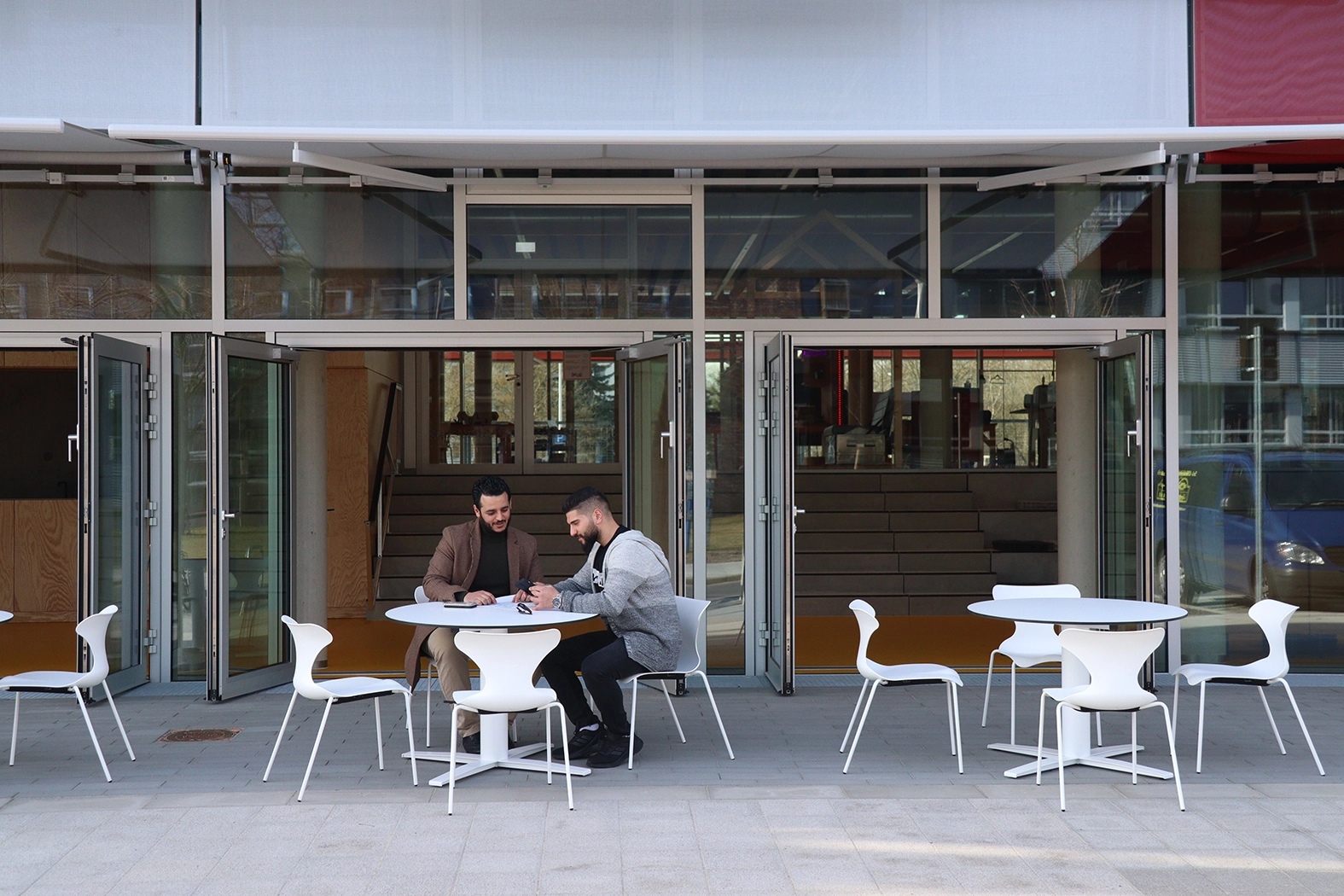 Ground-level view of a building pavilion showing open folding glass doors, with people sitting at minimalist white outdoor tables and chairs on a paved terrace.