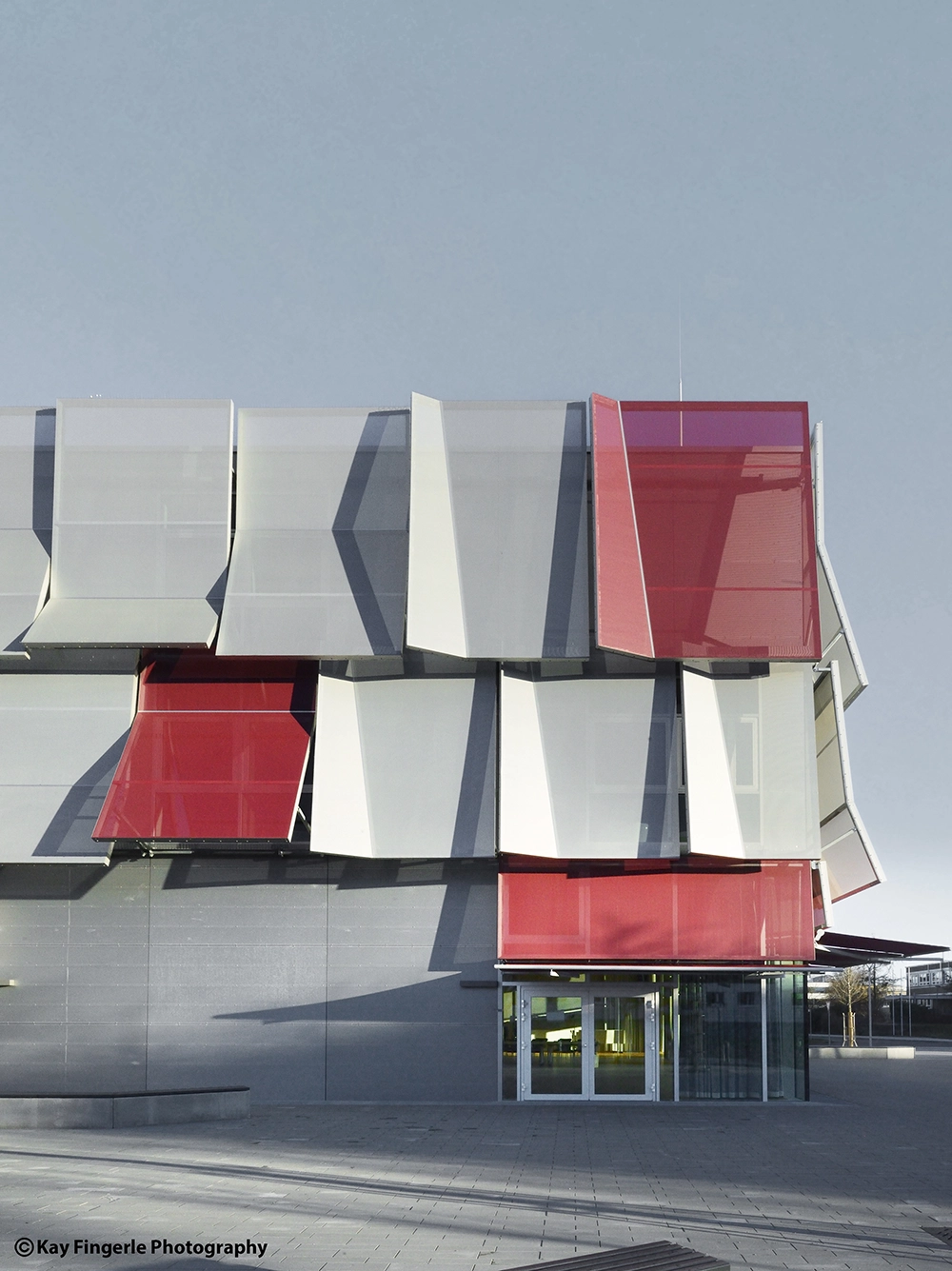 Frontal elevation crop of the institutional building facade, showcasing the overlapping rhythm of gray mesh screens and contrastingly vibrant red sunshades under a blue sky.