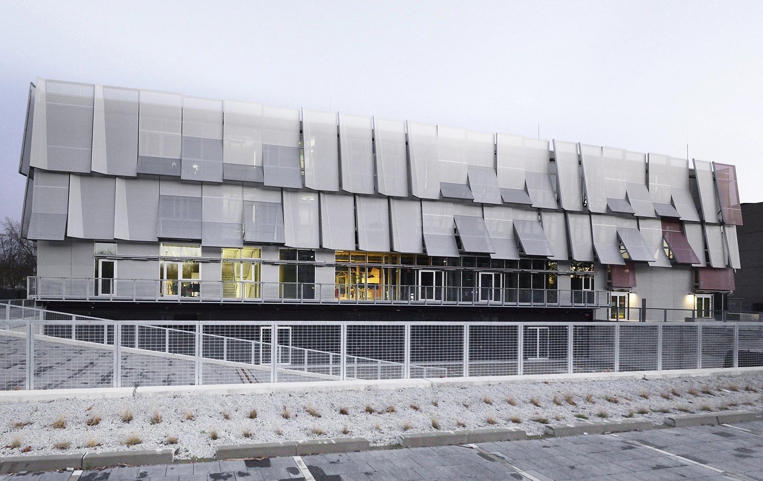 Wide exterior shot of a three-story institutional building wrapped in a dynamic, multi-layered skin of silver mesh panels and glass windows above a fenced perimeter.