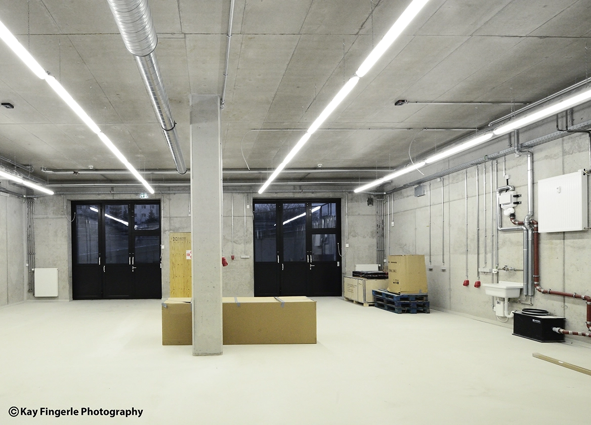 : Large industrial FabLab and workshop room with exposed concrete walls and ceiling, surface-mounted utility conduits, bright linear lighting strips, and black double doors.