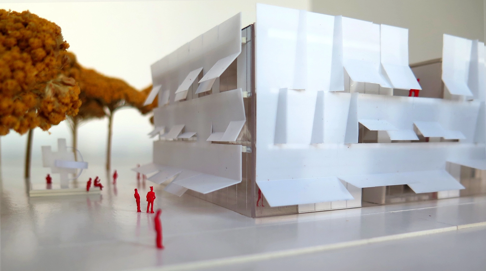 Architectural scale model of the building facade showing white translucent louvers at various angles of opening, with red miniature figures and dried yellow trees in the foreground.