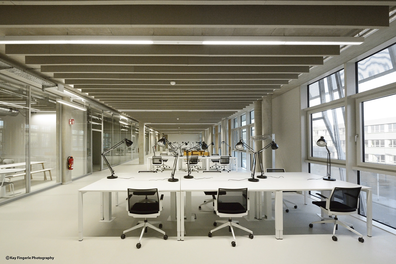 Modern open office work environment with long white desks, black swivel chairs, individual desk lamps, and linear acoustic baffles integrated into the concrete ceiling.