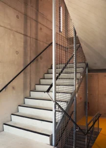 Interior utility staircase constructed from precast concrete steps, framed by industrial steel mesh safety balustrades against a smooth fair-faced concrete wall.