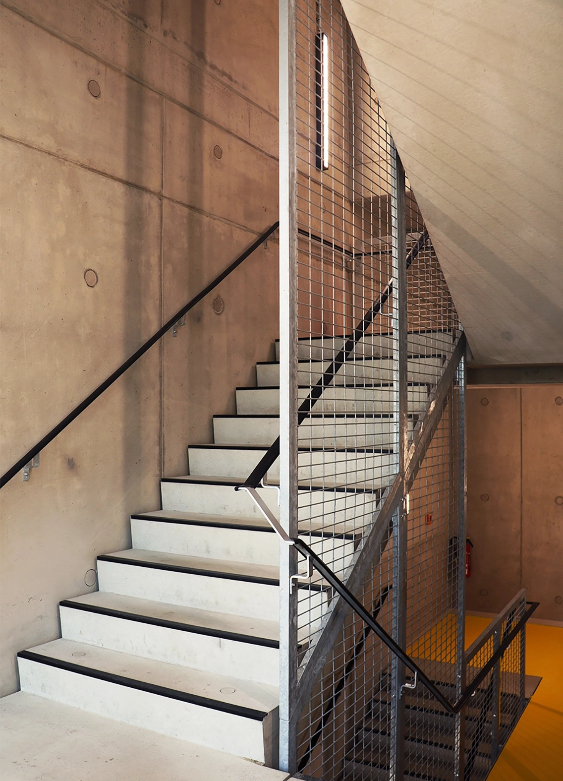 Interior utility staircase constructed from precast concrete steps, framed by industrial steel mesh safety balustrades against a smooth fair-faced concrete wall.